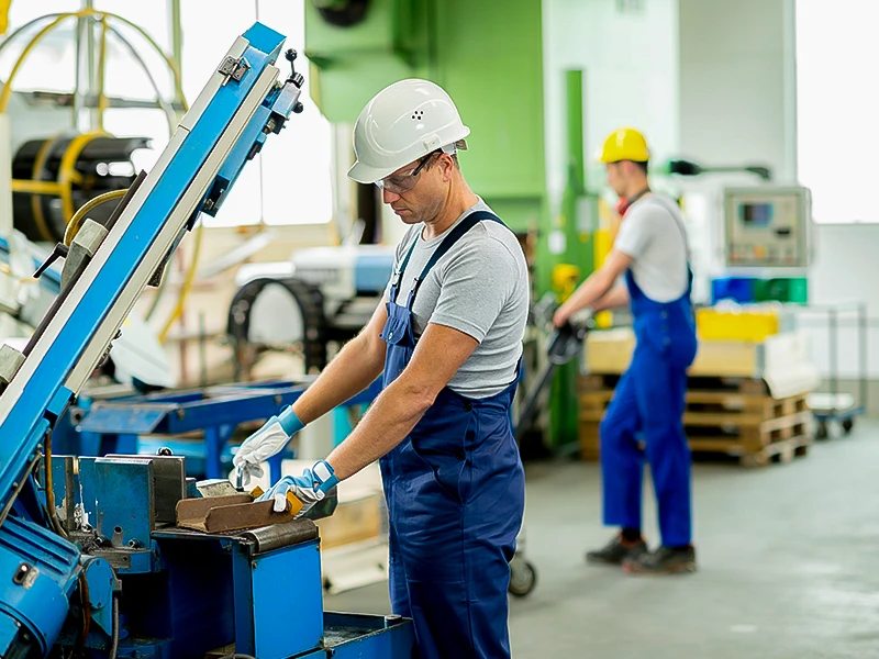 The man in the foreground is working on a machine and the man in the background is pulling a pallet truck.