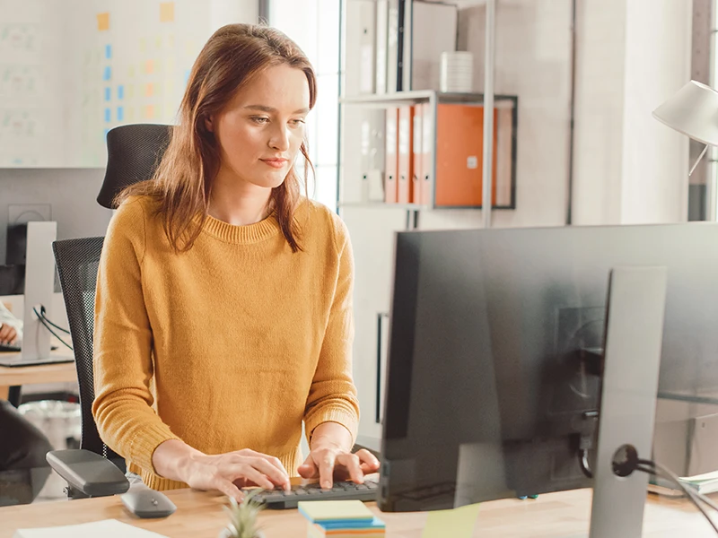 Eine junge Frau mit gelbem Pullover arbeitet am Computer-Bildschirm.