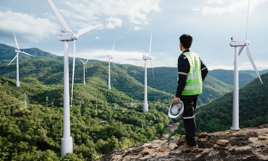 A person in work clothes looks out over an idyllic valley full of wind turbines.