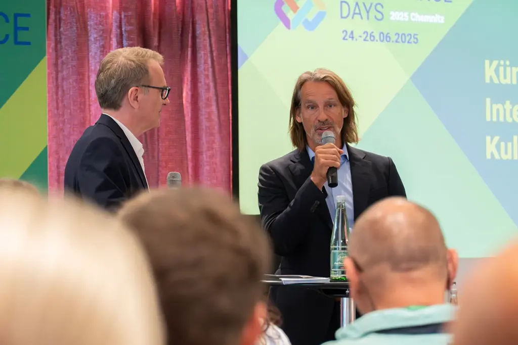 Zwei Männer halten eine Präsentation auf einer Bühne. Hinter ihnen ist das Bild eines Projektors sichtbar.