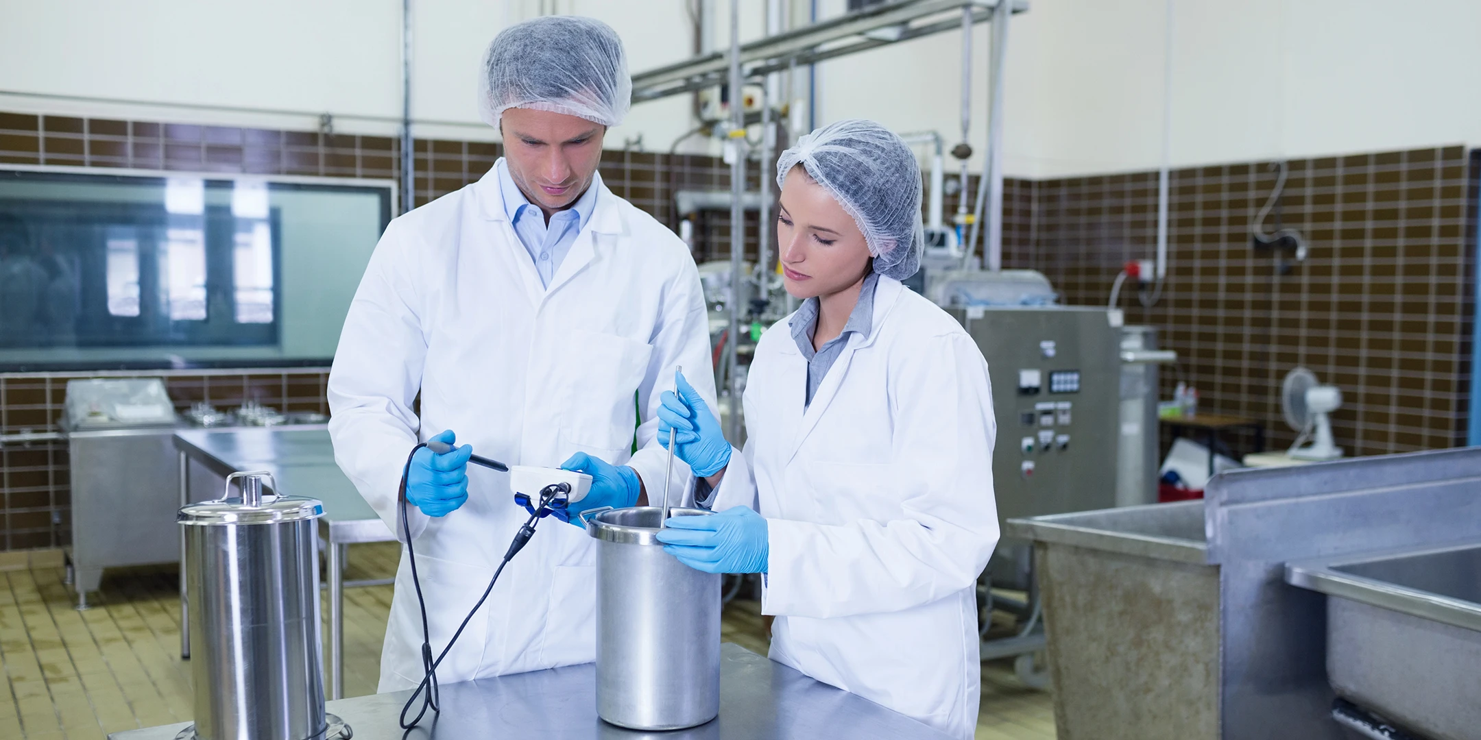 Zwei Mitarbeitende in Schutzkleidung untersuchen eine Lebensmittel-Probe nach HACCP