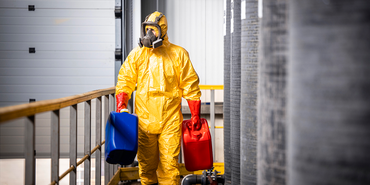 A person in a protective suit carries two canisters containing hazardous materials.