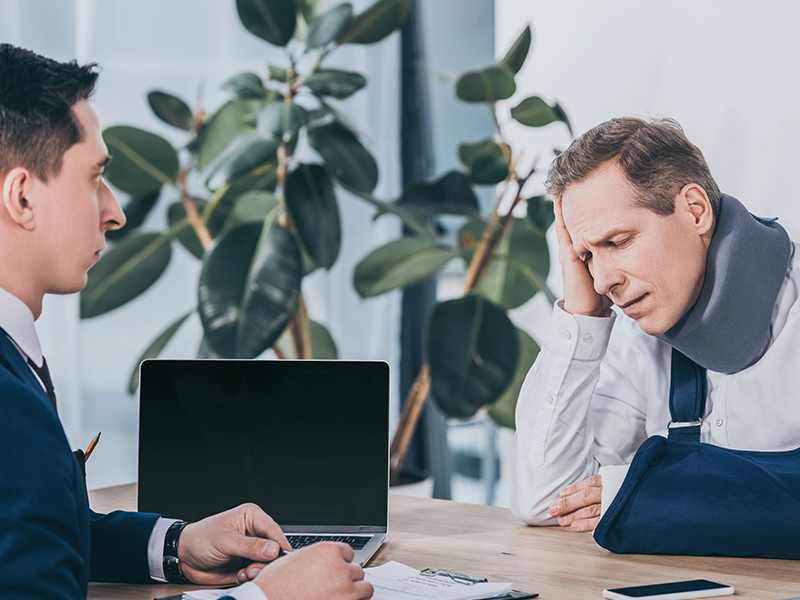 Zwei Männer sitzen sich gegenüber. Ein Mann hält sich den Kopf und der andere sitzt von seine, Laptop.