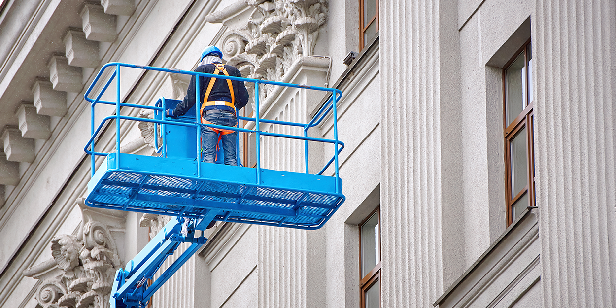 Ein Arbeiter steht auf einer hochgefahrenen Hebebühne an einer weißen Fassade