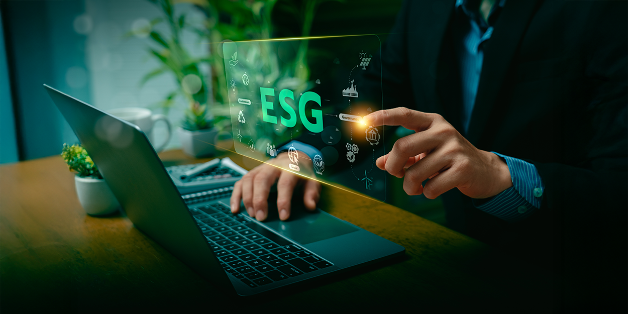 A man is working on a laptop and tapping his finger on a hologram with ESG symbols.